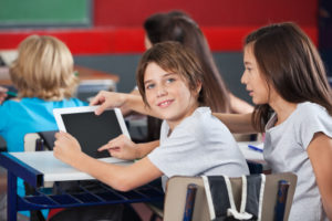 Young boy uses tablet with his peers in his classroom while smiling.