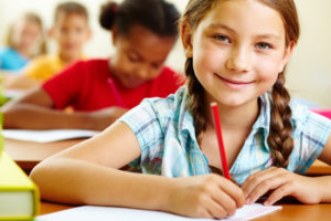 Young girl with hair in braids smiles while writing on her paper in her classroom.