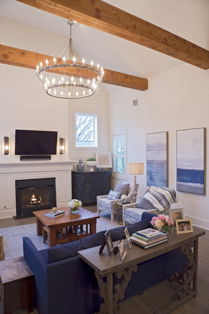  Stained pine beams and a metal two-tier chandelier soar above the living room where a black honed granite fireplace with custom millwork mantle bring warmth to the transitional setting. A pair of tall distressed black Demilune consoles bookmark the fireplace providing storage. Blue velvet upholstered sofa and blue and white patterned fabric on the washed wooden legged arm stairs give the room its casual, coastal vibe. 